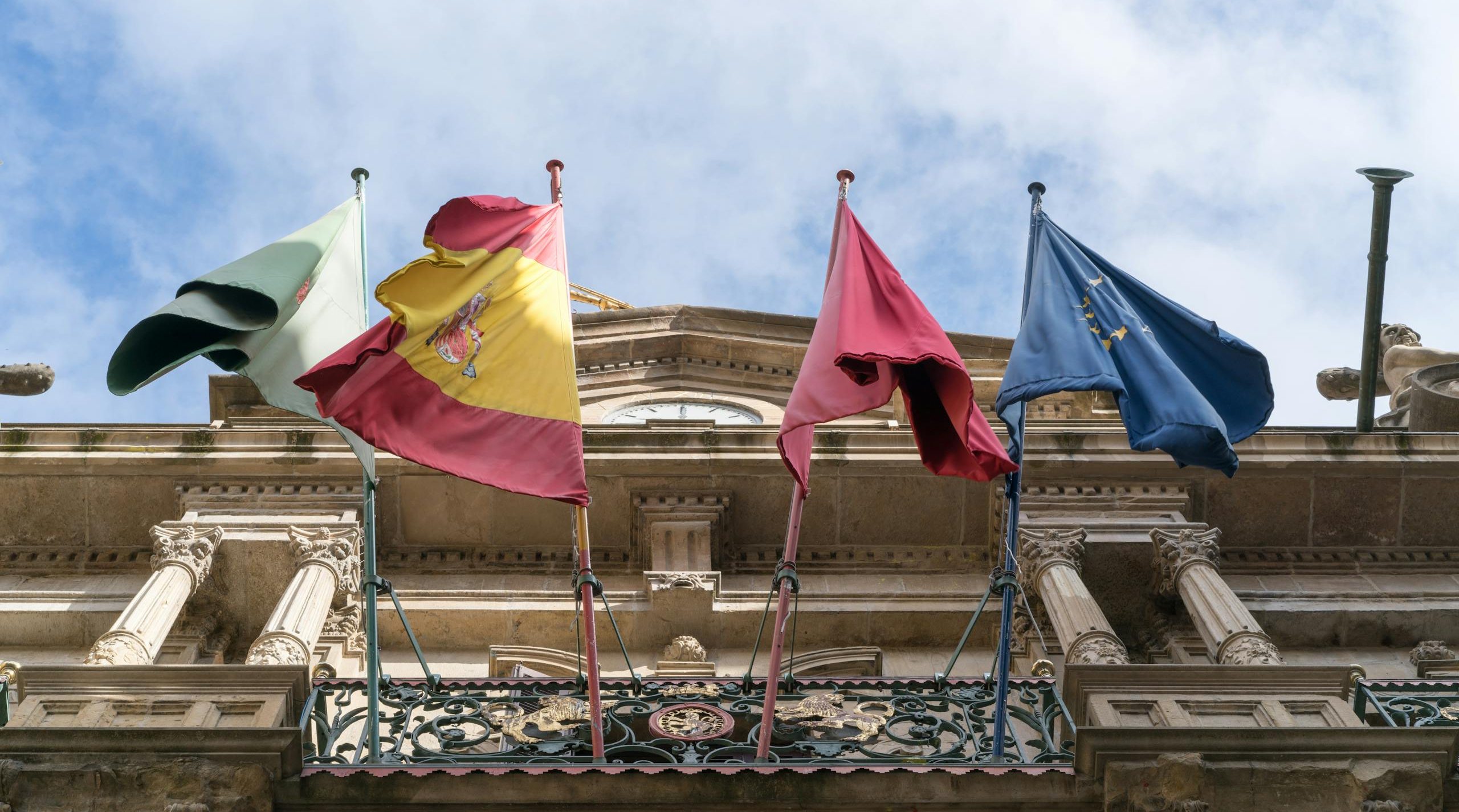 Flags of Spain, Italy, and EU on a historic building facade, symbolizing international unity.